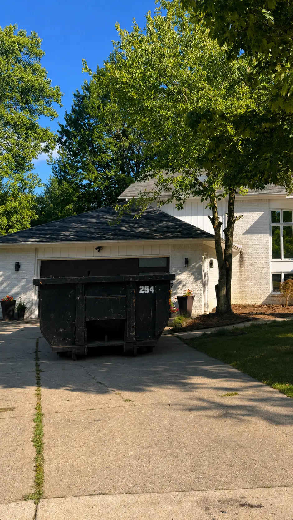 Black roll-off dumpster rental parked in a residential driveway at a white brick home in West Bloomfield, Michigan